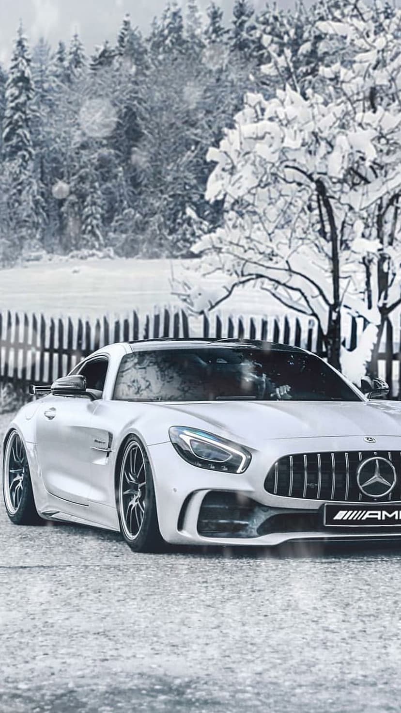White Mercedes-AMG parked on a wet snowy road with falling flakes and a rustic wooden fence in a winter forest.