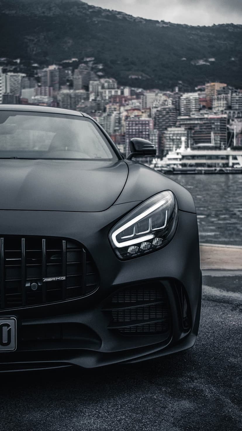 Matte black Mercedes-AMG GT R parked on a waterfront with a city skyline and distant mountains behind it.