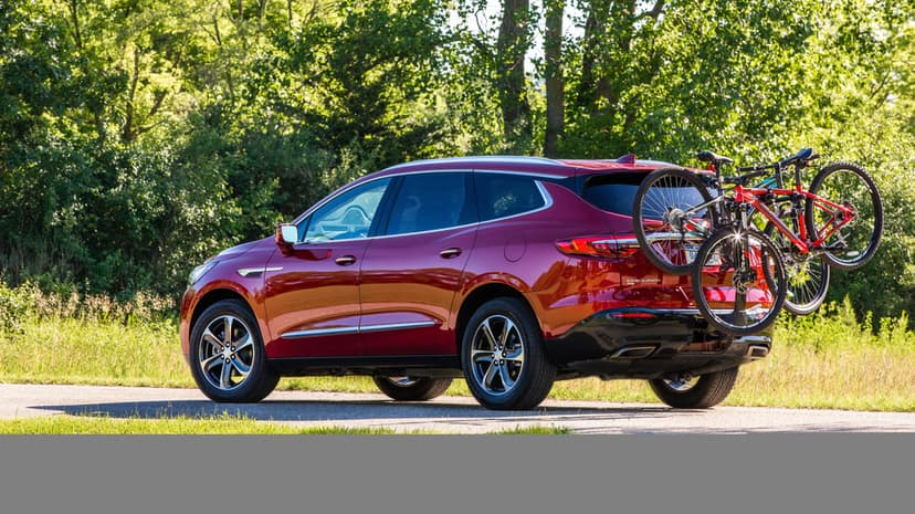 Red SUV with a bike rack on a paved road beside a lush green forest under bright summer daylight.