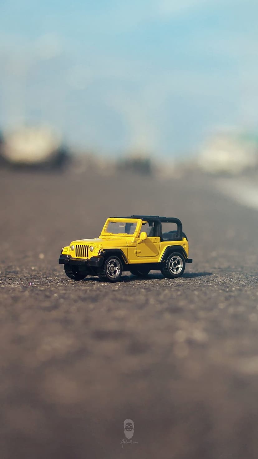 Yellow miniature Jeep Wrangler with a black top sits on a dark asphalt road under bright daylight.
