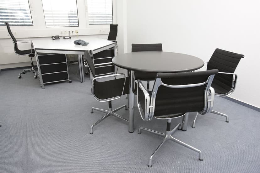 Black mesh executive chairs surround a circular conference table in a modern office with large window blinds.