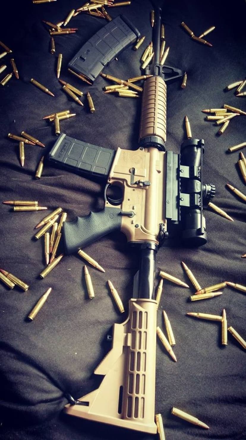 Tan assault rifle with optical scope rests on a textured dark surface among scattered metallic cartridges.