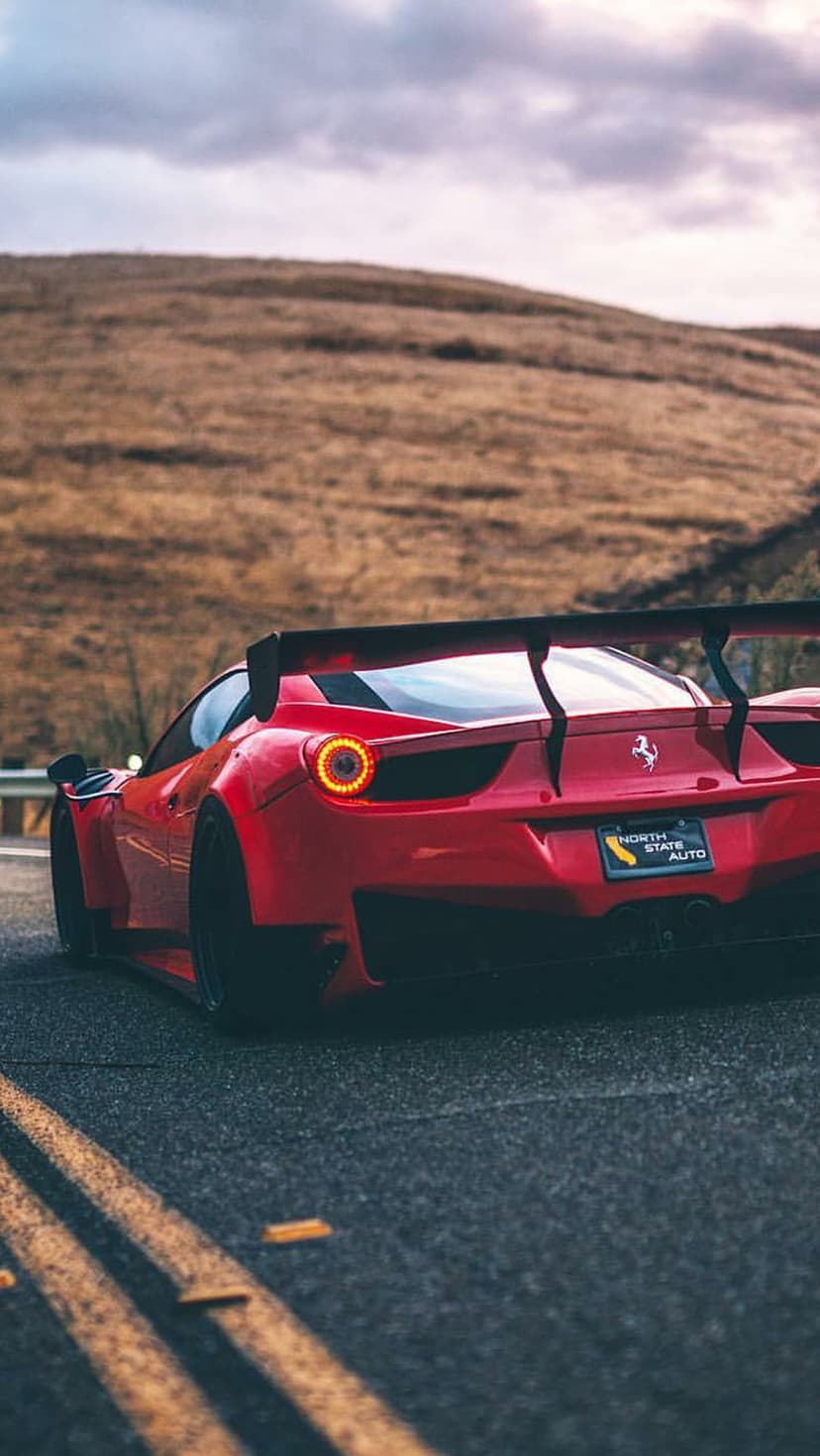 Red Ferrari with glowing taillights and a rear wing drives on a paved road bordered by golden grassy hills.
