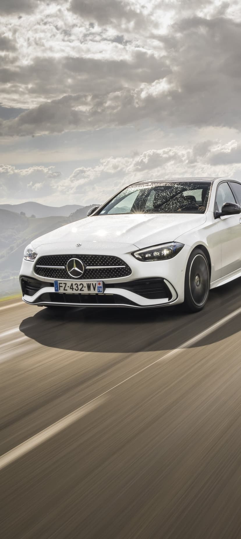 White Mercedes-Benz sedan driving on a blurred road with a dramatic cloudy sky and rolling hills background.