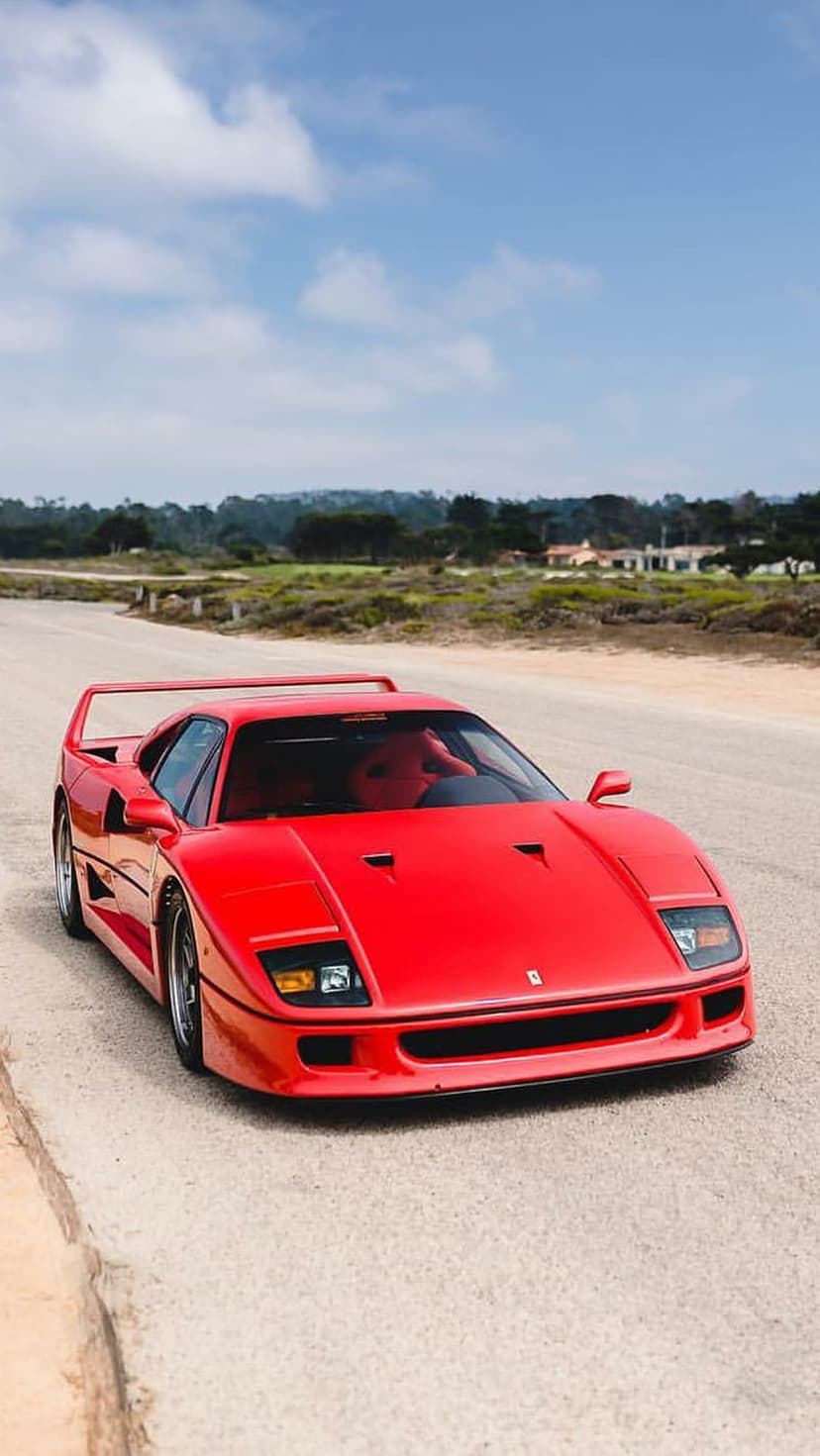 Red Ferrari F40 parked on a dusty road with green trees and a cloudy blue sky in a vertical orientation.