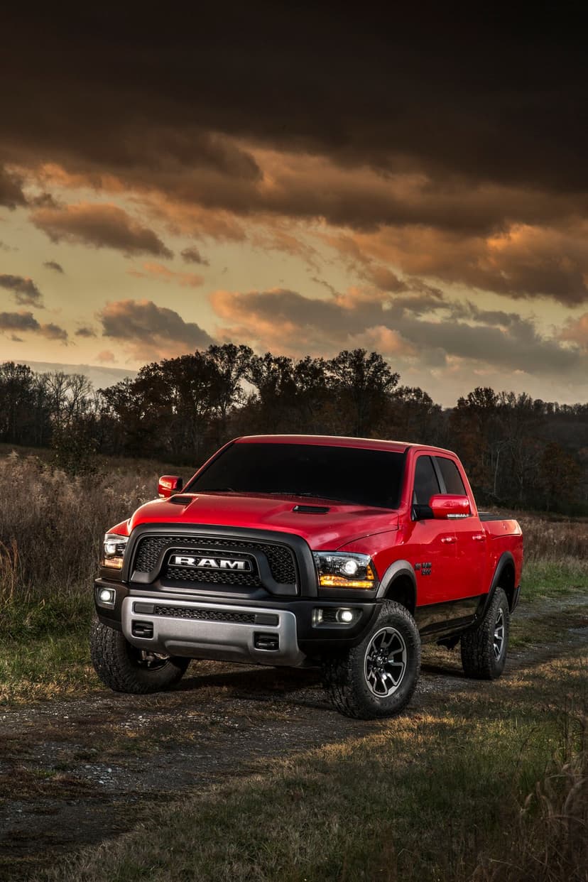 Red Ram 1500 Rebel truck with glowing headlights parked on a dirt trail under a dramatic golden sunset sky.