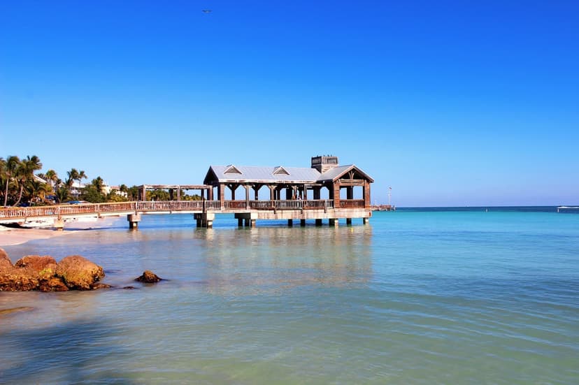 Wooden pier with a pavilion extends over clear turquoise water near palm trees on a sunny Key West beach.
