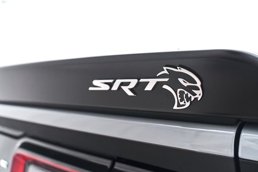 Silver SRT Hellcat emblem and snarling cat logo mounted on a matte black rear car spoiler under bright light.