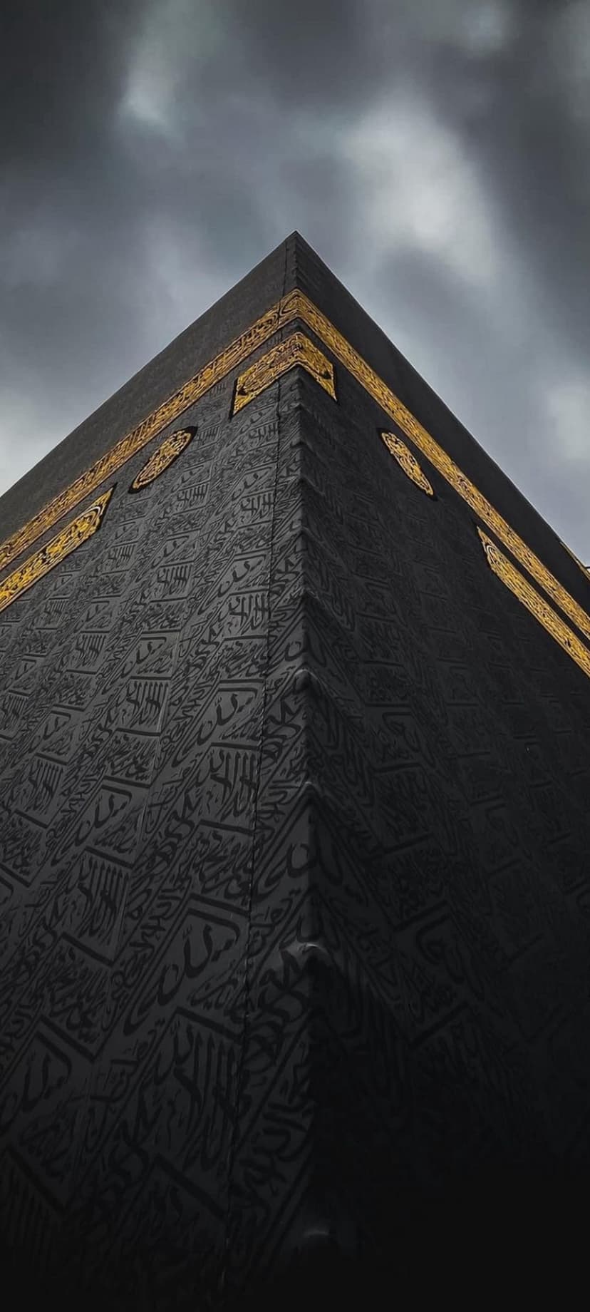 Kaaba corner draped in black silk with gold Arabic calligraphy against a dark, cloudy sky in a vertical view.