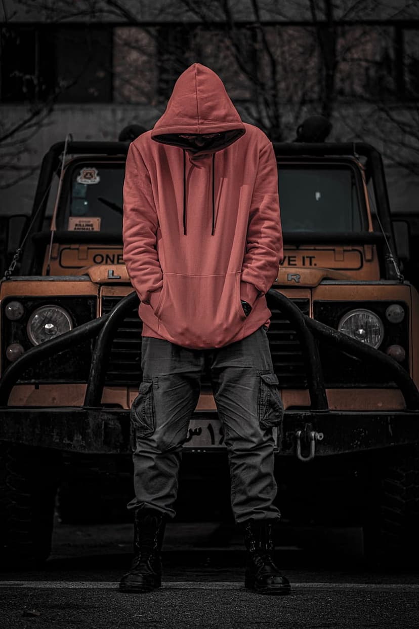 Man wearing a vibrant orange hoodie and cargo boots stands beside a yellow off-road SUV in dark lighting.