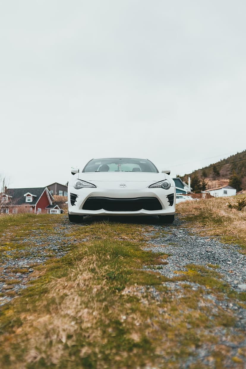 White sports car parked centrally on a gravel driveway with dry grass and houses under an overcast gray sky.
