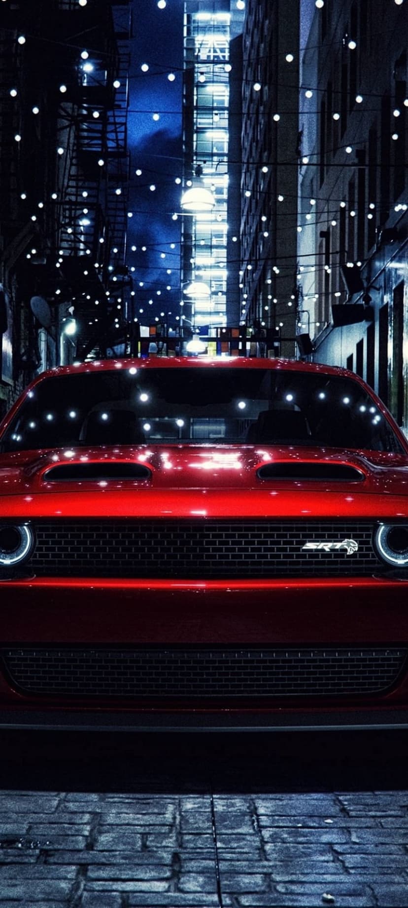 Red Dodge Challenger parked on wet cobblestones at night under twinkling string lights in a narrow urban alley.