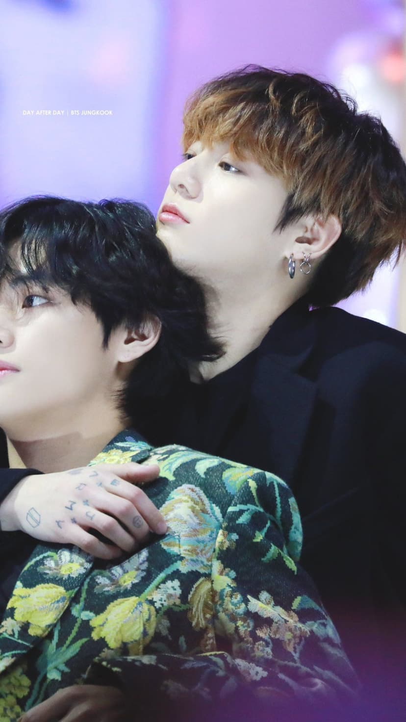 Taehyung wraps his arms around Jungkook’s shoulders from behind while leaning his head in a soft bokeh background.