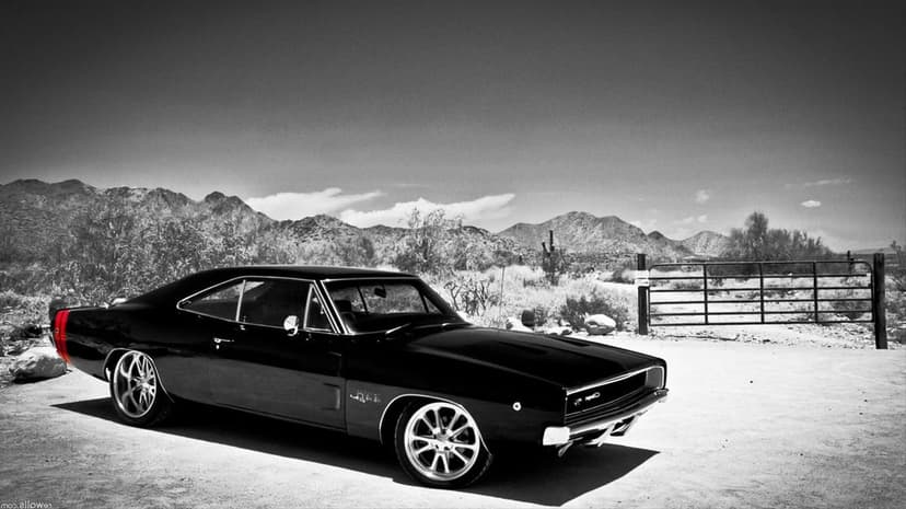 Black Dodge Charger muscle car with a red stripe parked on a dirt path in a rugged desert mountain landscape.