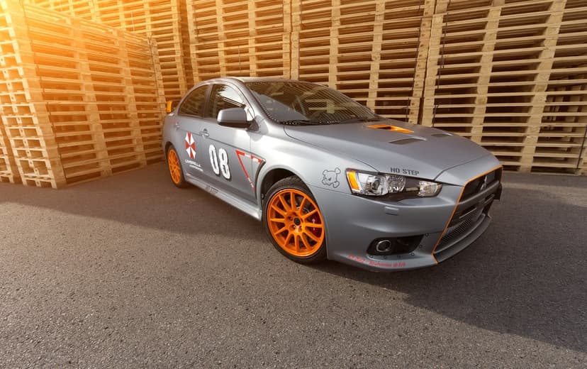 Matte grey Mitsubishi Lancer with orange wheels and Umbrella decals by wooden pallets under golden sunset flare.