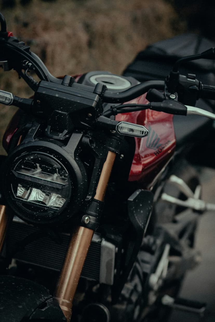 Red Honda motorcycle headlight and bronze front forks covered in water droplets against a blurred background.