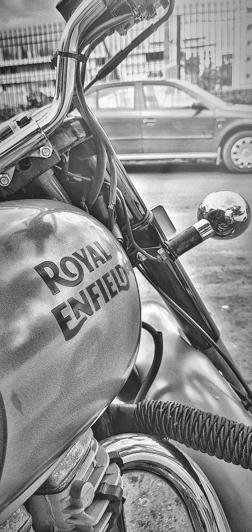 Royal Enfield motorcycle fuel tank features a chrome logo and handlebars within a blurred monochrome city street.