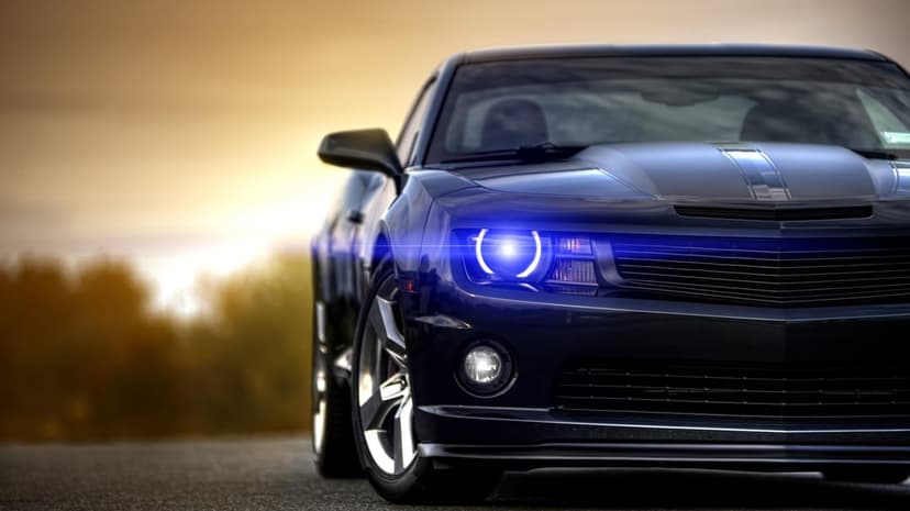 Front view of a dark blue muscle car with a glowing blue halo headlight against a sunset background.
