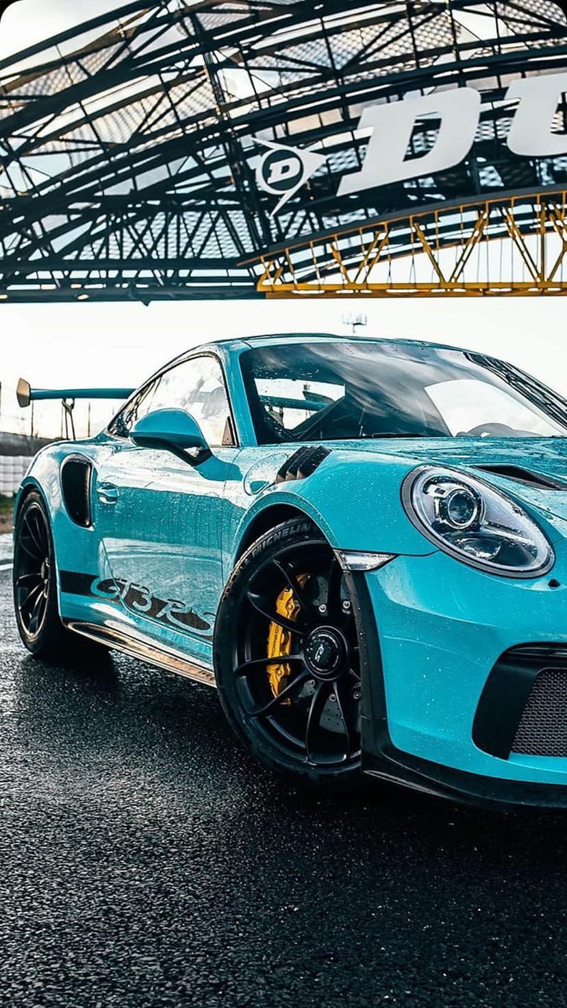 Blue Porsche GT3 RS on a wet asphalt track with yellow brake calipers and the Dunlop bridge in the background.