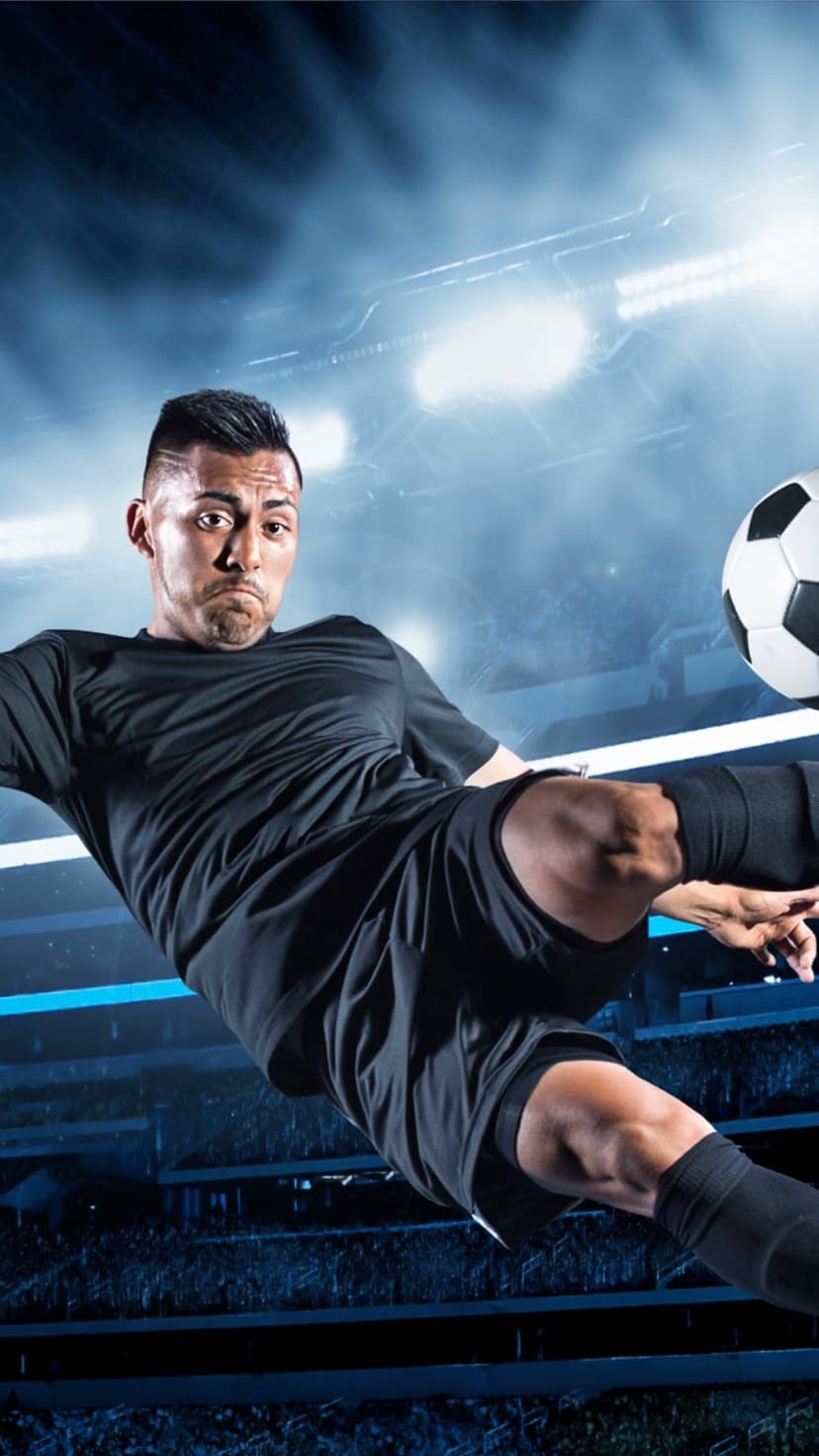 Soccer player in a black kit kicks a ball mid-air against bright stadium floodlights during a night game.