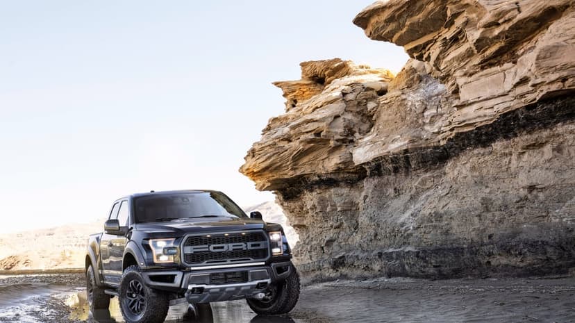 Black Ford Raptor pickup truck parked next to a layered desert cliff under a bright, clear sky background.