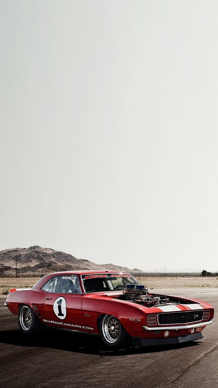 Red 1969 Chevrolet Camaro drag car with a blower hood scoop on a tarmac track against desert mountains.