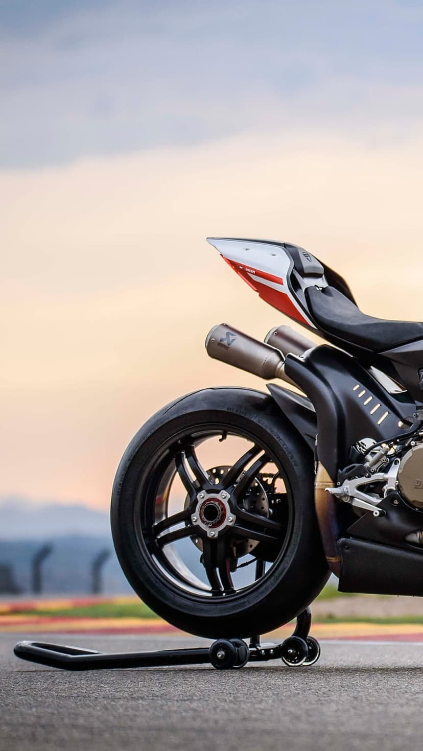 Black Ducati motorcycle rear wheel and dual exhaust pipes on a track stand with a blurred sunset background.