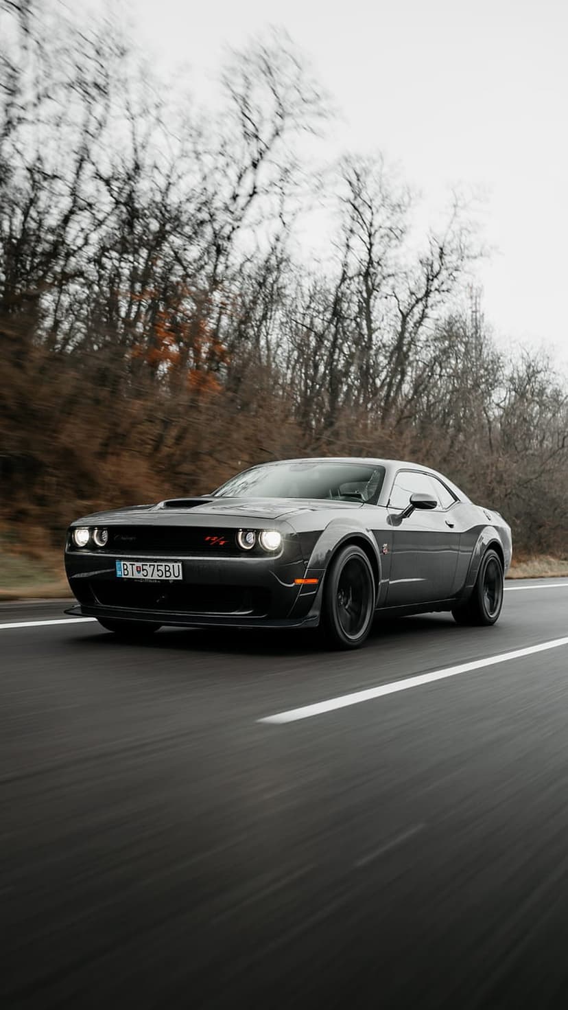 Dark gray Dodge Challenger muscle car driving fast on a paved road with blurred winter trees and cloudy sky.