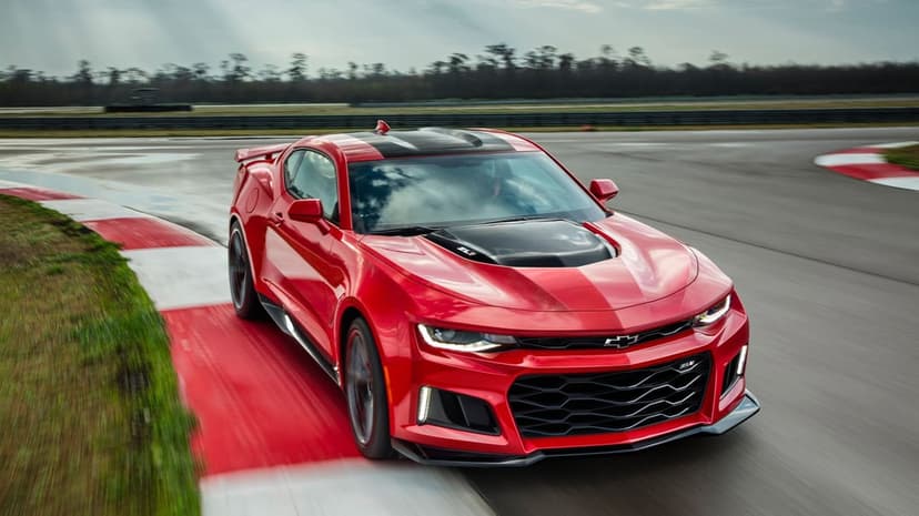 Red Chevrolet Camaro ZL1 racing on an asphalt track with motion blur along red and white curbs under overcast skies.