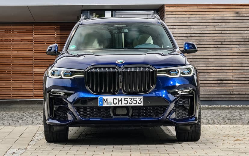 Front view of a dark blue BMW X7 luxury SUV with black grilles parked on a stone driveway by a modern wood building.