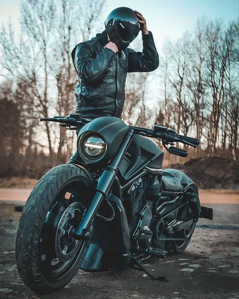 Biker in a leather jacket and helmet adjusts his gear beside a black motorcycle against a backdrop of trees.