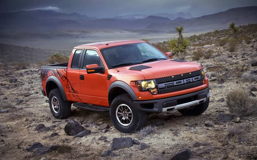 Orange Ford F-150 SVT Raptor truck parked on rocky desert ground with mountains under a cloudy sky.