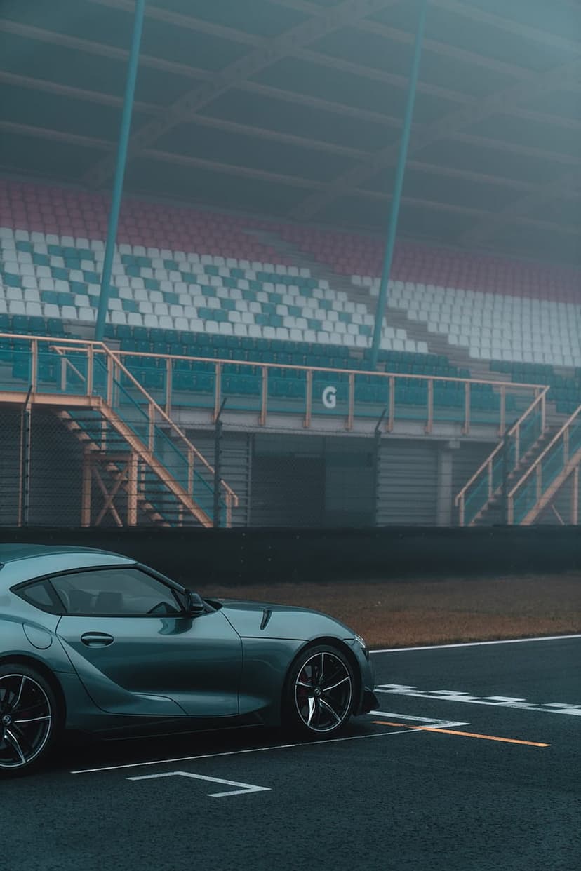 Toyota Supra sports car parked on a wet asphalt racetrack with stadium seating blurred in the misty background.
