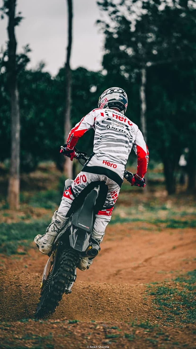 Motocross rider in a red and white suit leans a motorcycle into a dusty turn on a wooded off-road racing track.