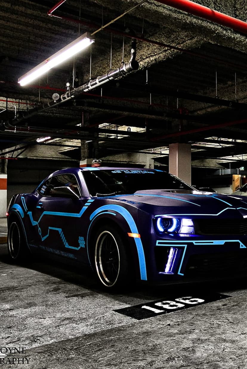 Dark purple Chevrolet Camaro with blue glowing circuitry decals and bright headlights in a dim concrete garage.