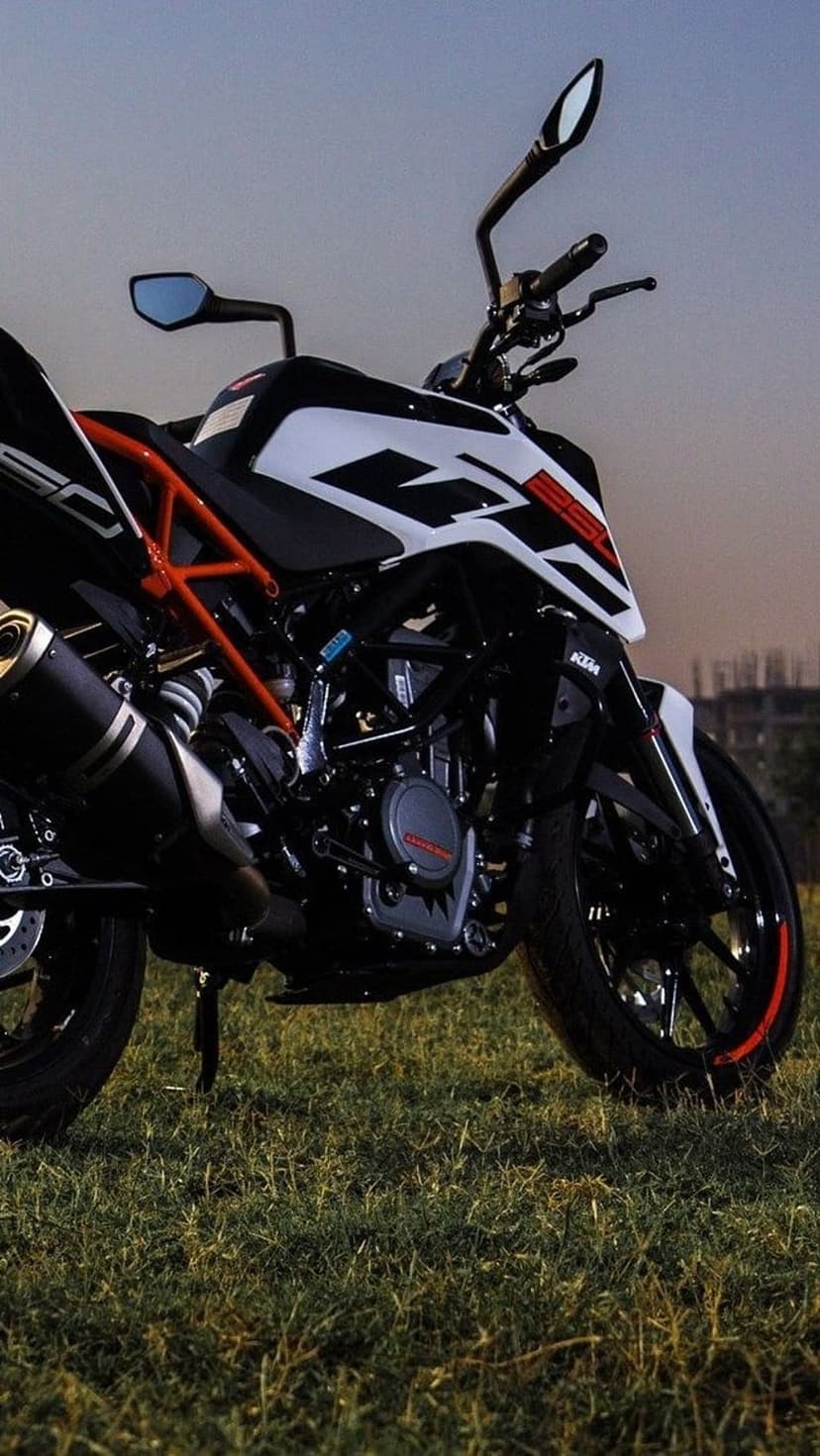 KTM motorcycle on green grass with a sunset sky, showing the orange frame, black engine, and chrome exhaust pipe.