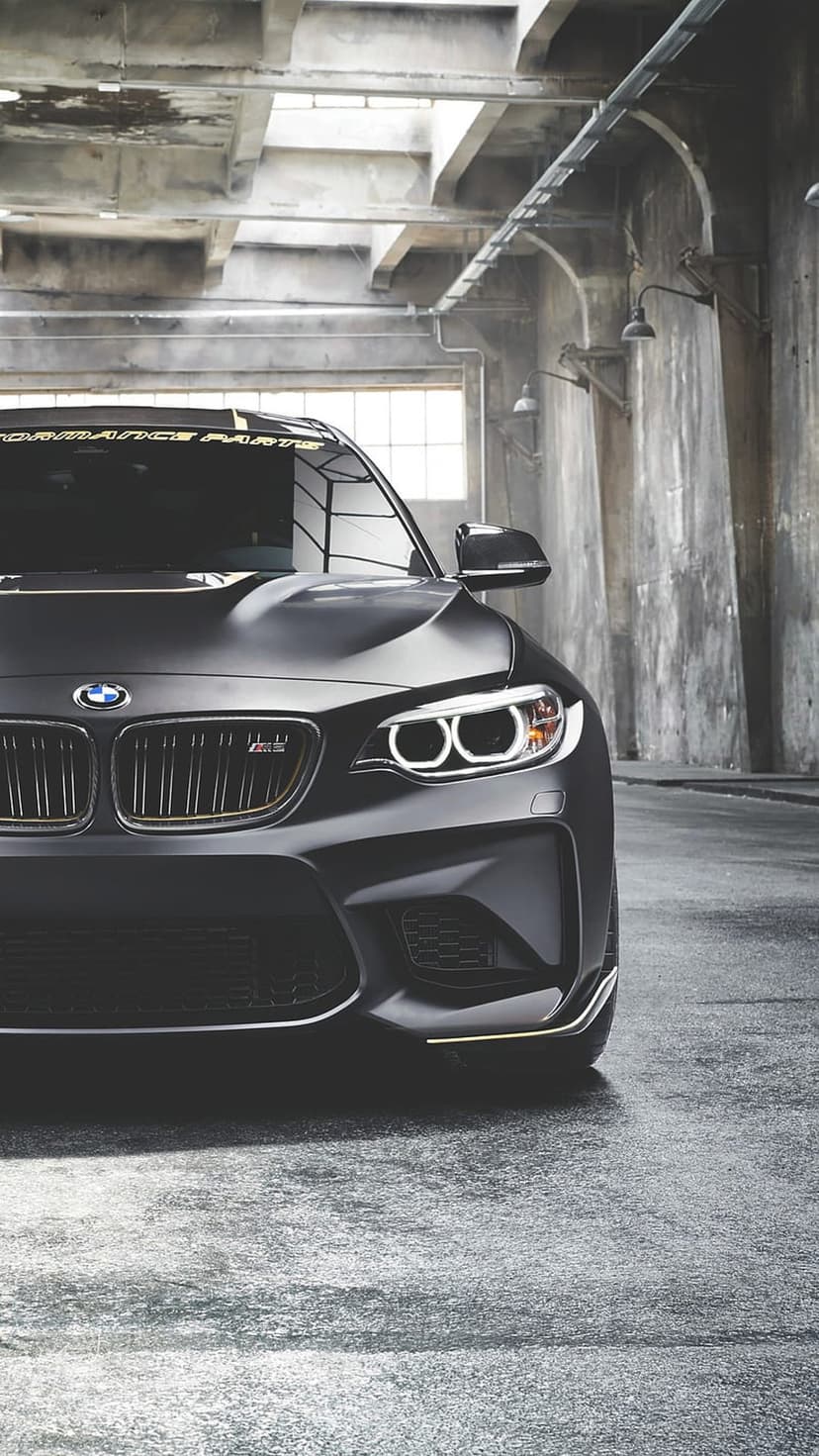 BMW M2 with a matte black finish and glowing halo headlights parked on a wet floor in a dark industrial garage.