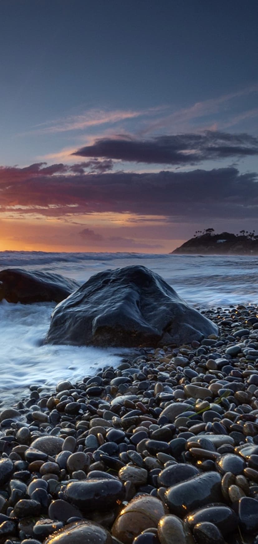 Dark jagged rocks sit amidst misty ocean waves under a vibrant orange and deep purple twilight sky at a beach.