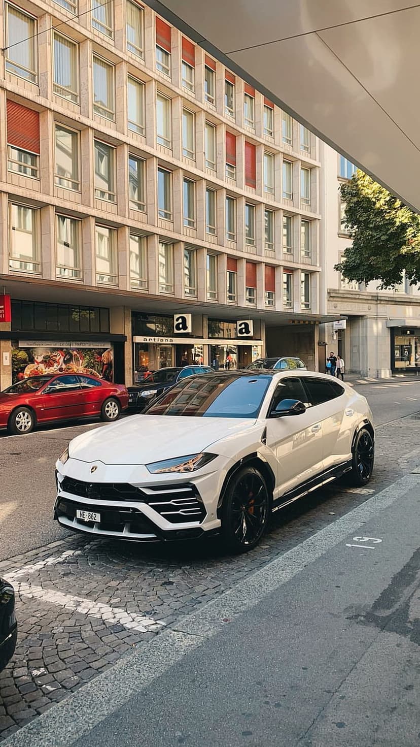 White Lamborghini Urus SUV parked on a gray cobblestone road in front of a modern building with red awnings.