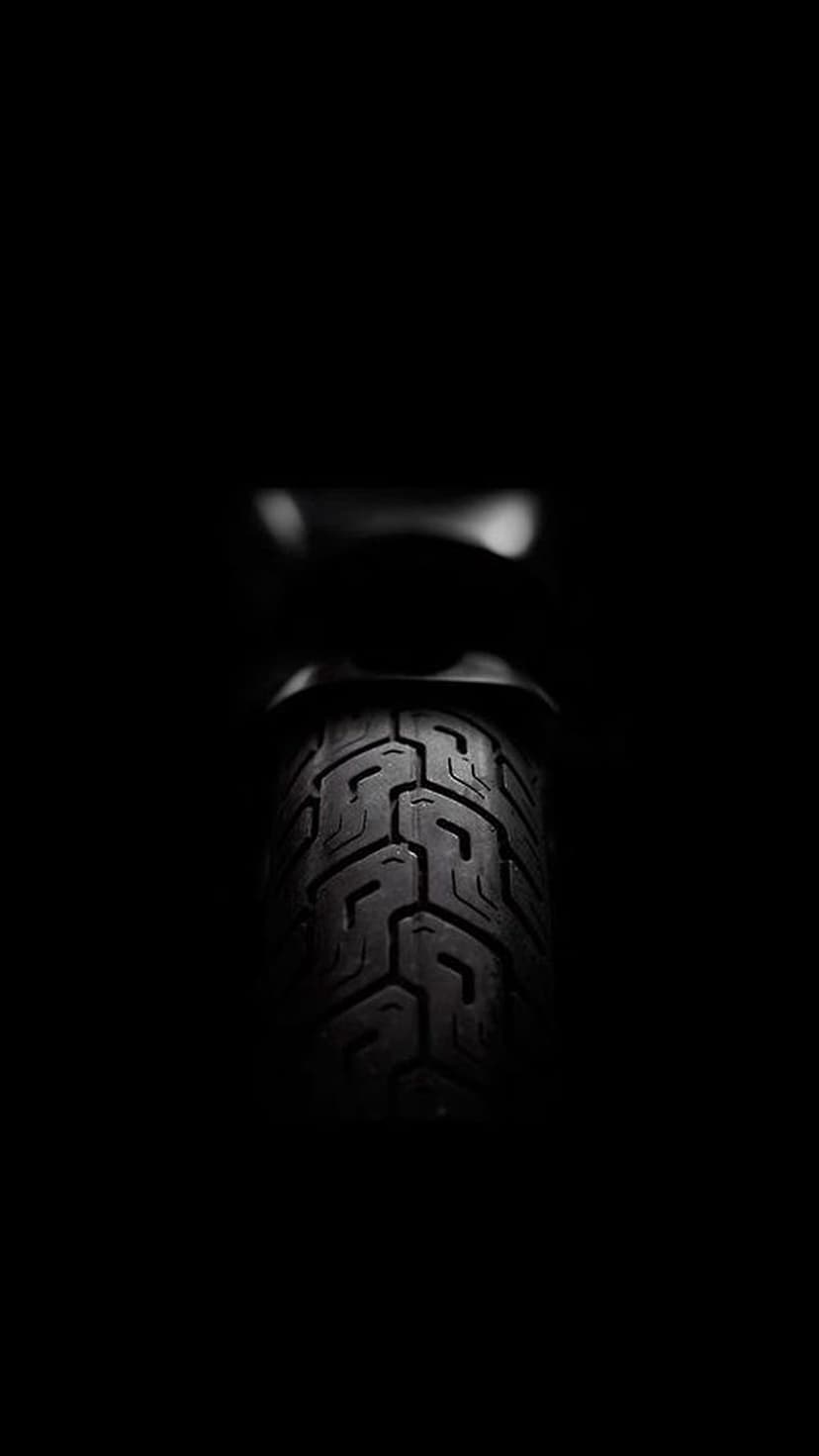 Motorcycle tire tread with deep rubber grooves and sharp high-contrast lighting against a pitch black background.