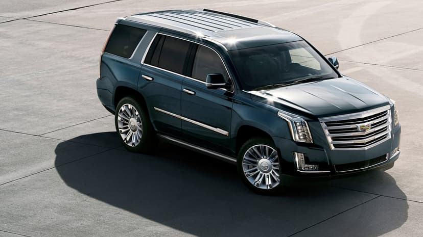 Dark teal Cadillac Escalade SUV with chrome wheels parked on a flat concrete surface with a long shadow.