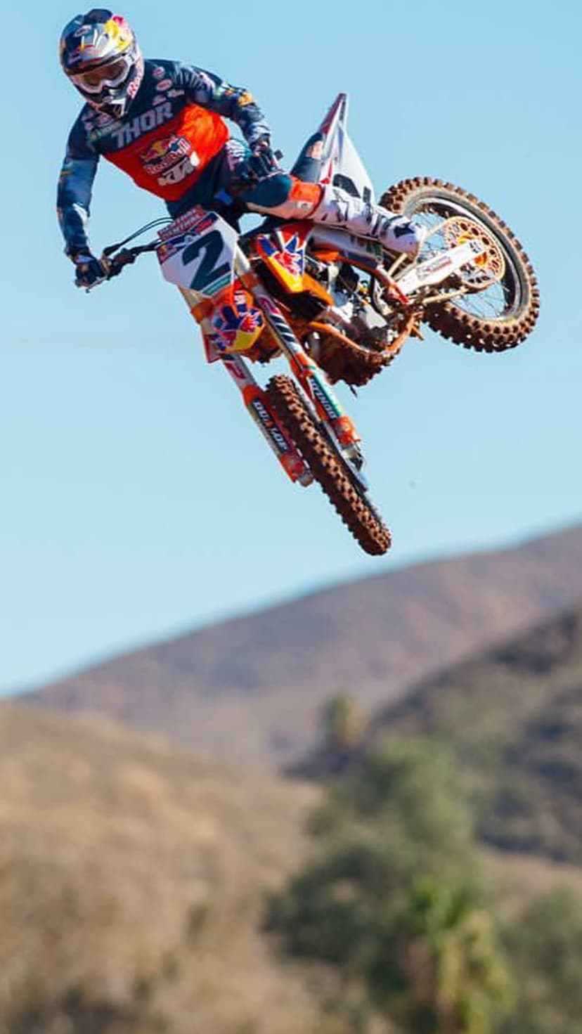 Motocross rider in red and blue gear jumps an orange KTM bike high above a blurred green and brown landscape.