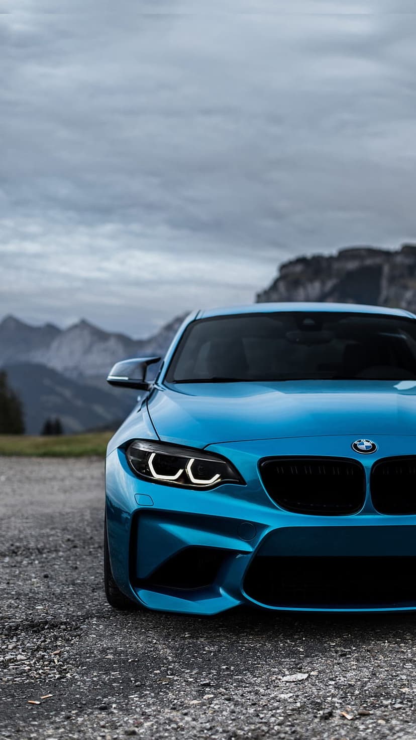 Blue BMW M2 sports car parked on a gravel trail with jagged mountains and a cloudy sky in the background.