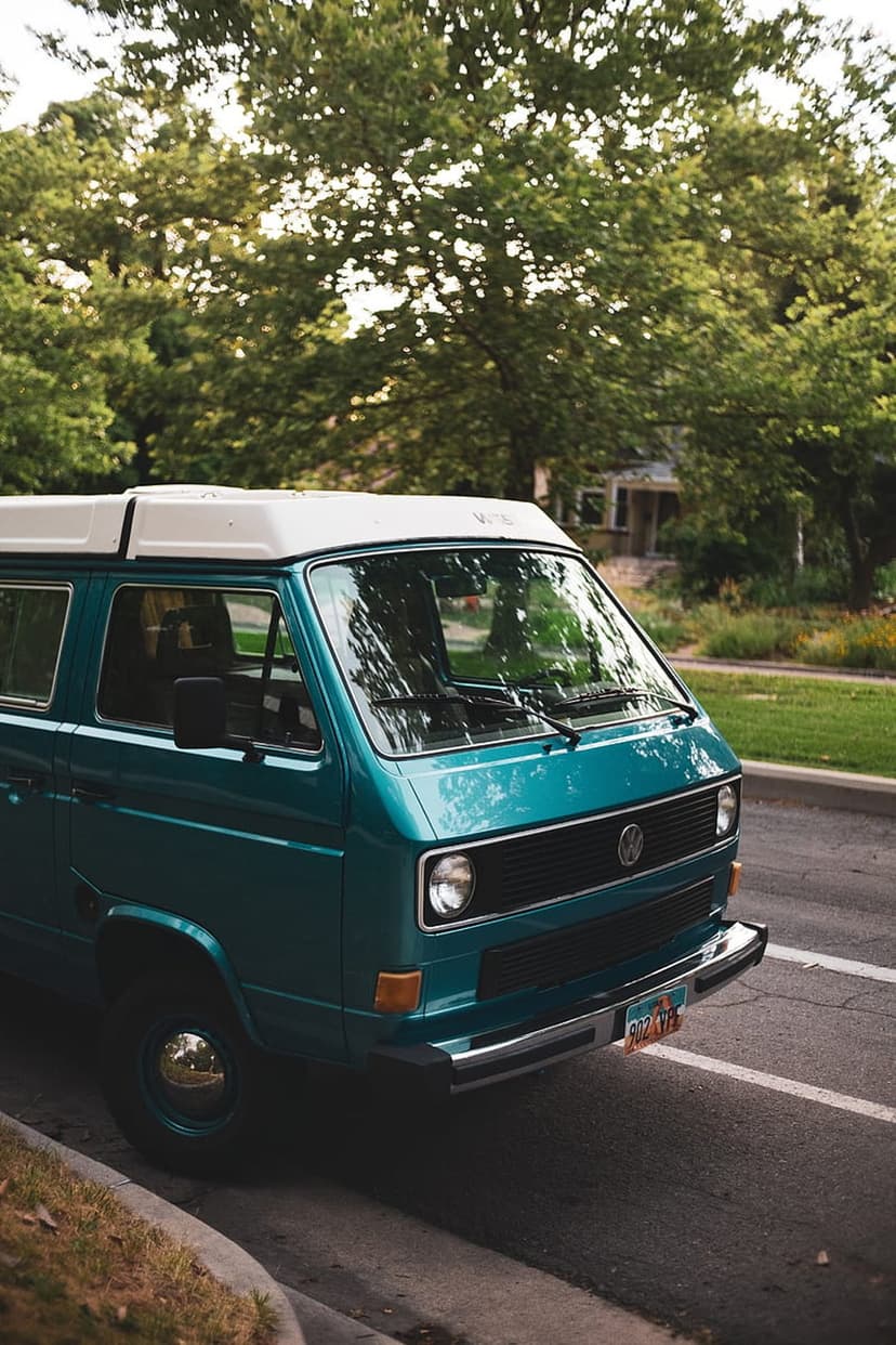 Teal Volkswagen camper van with white pop-top parked on a sun-dappled street under lush green trees.