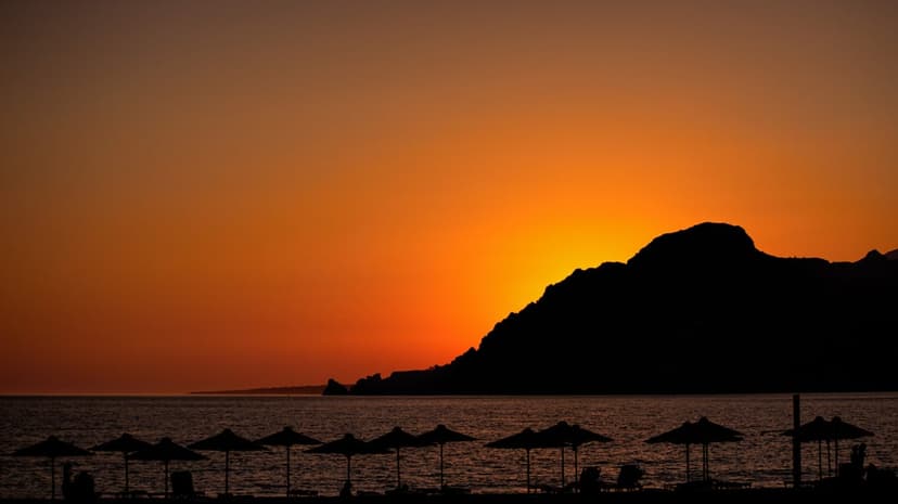 Beach umbrellas and lounge chairs silhouetted against a mountain sunset and orange sea on a tropical shoreline.