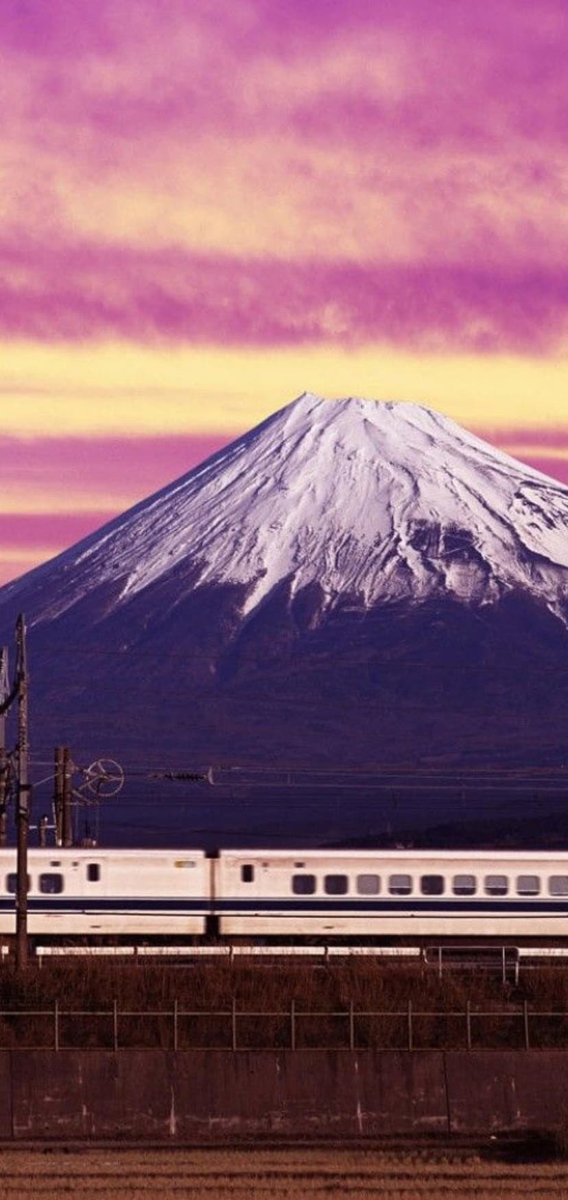 White bullet train speeds past snow-capped Mount Fuji under a vibrant pink and orange sunset cloudscape.