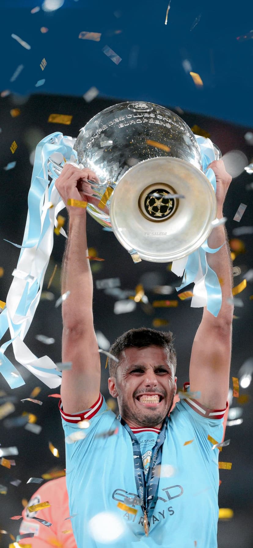 Manchester City player holding the Champions League trophy overhead with falling silver confetti and streamers.