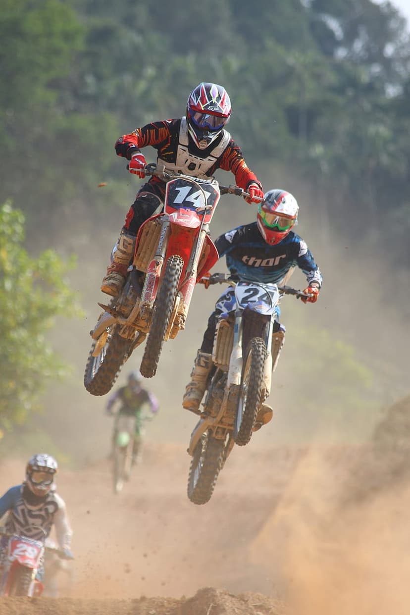 Two motocross riders catch air on dirt bikes with dust clouds trailing behind against a blurred green forest.