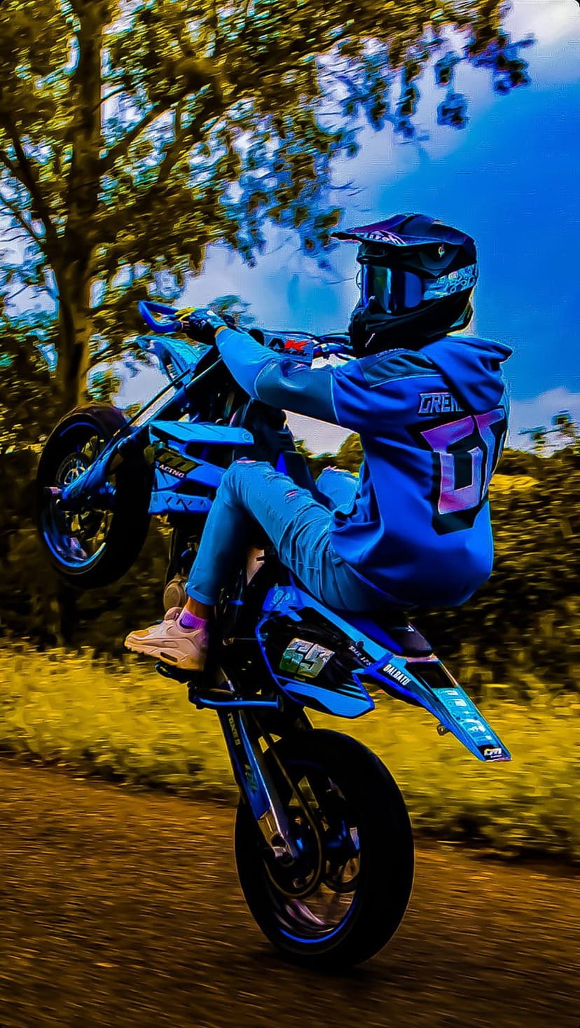 Motocross rider in blue gear performs a wheelie on a dirt path with dust clouds and green trees in the background.