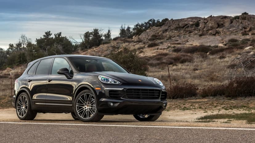 Black Porsche Cayenne SUV parked on a roadside with rocky hills and a cloudy sky in the background.